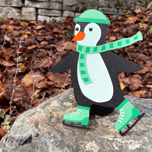 Paper penguin character wearing a green hat and scarf ice skating on a rock, part of the Waddle & Whirl collection by Michelle Ward Designs.