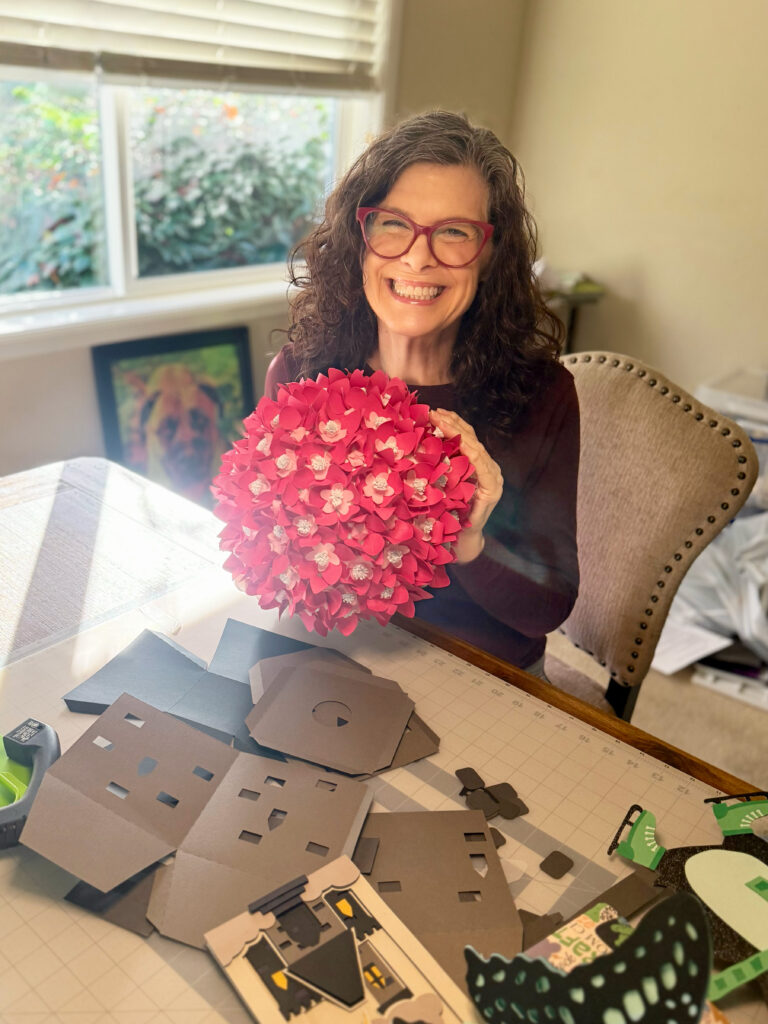 Michelle Ward smiling at her craft table, holding a handmade paper hydrangea sphere, surrounded by colorful 3D paper pieces and creative tools.