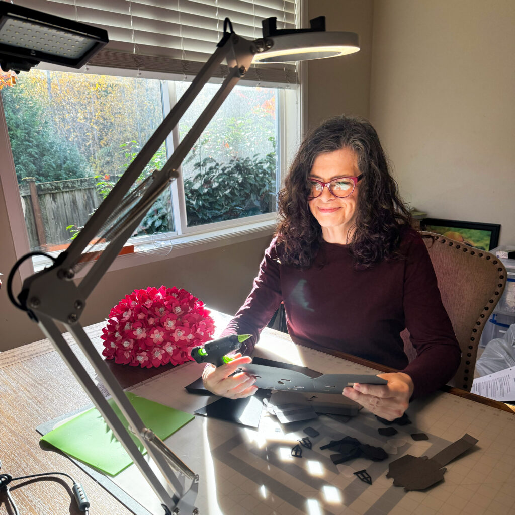 Michelle Ward working at her craft table with a glue gun and paper pieces scattered under a bright lamp.
