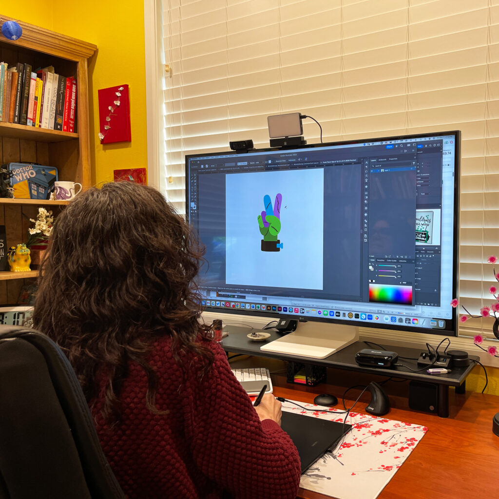 Michelle Ward working in Adobe Illustrator on a large screen, creating a colorful stitched peace sign design in her home studio.