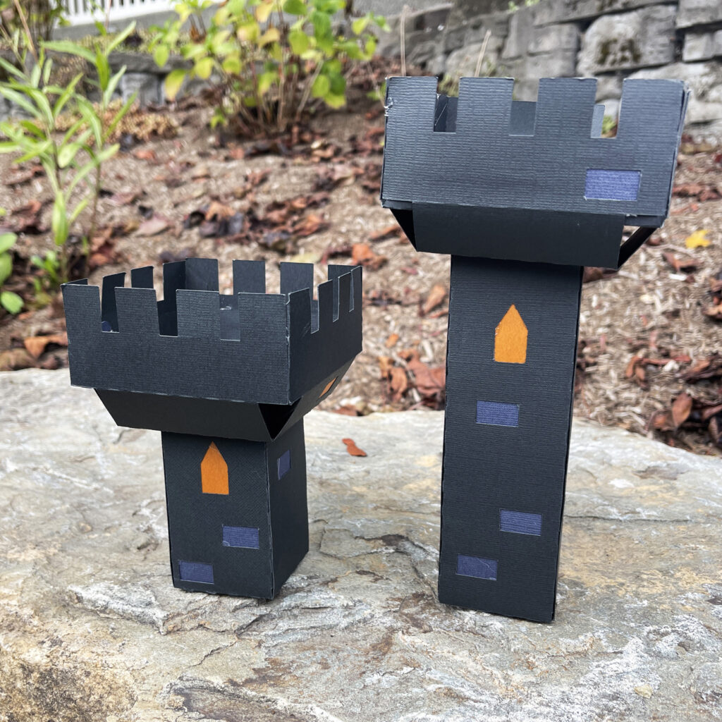 Two 3D paper castle battlement towers in black and gray cardstock sitting on a rock outdoors