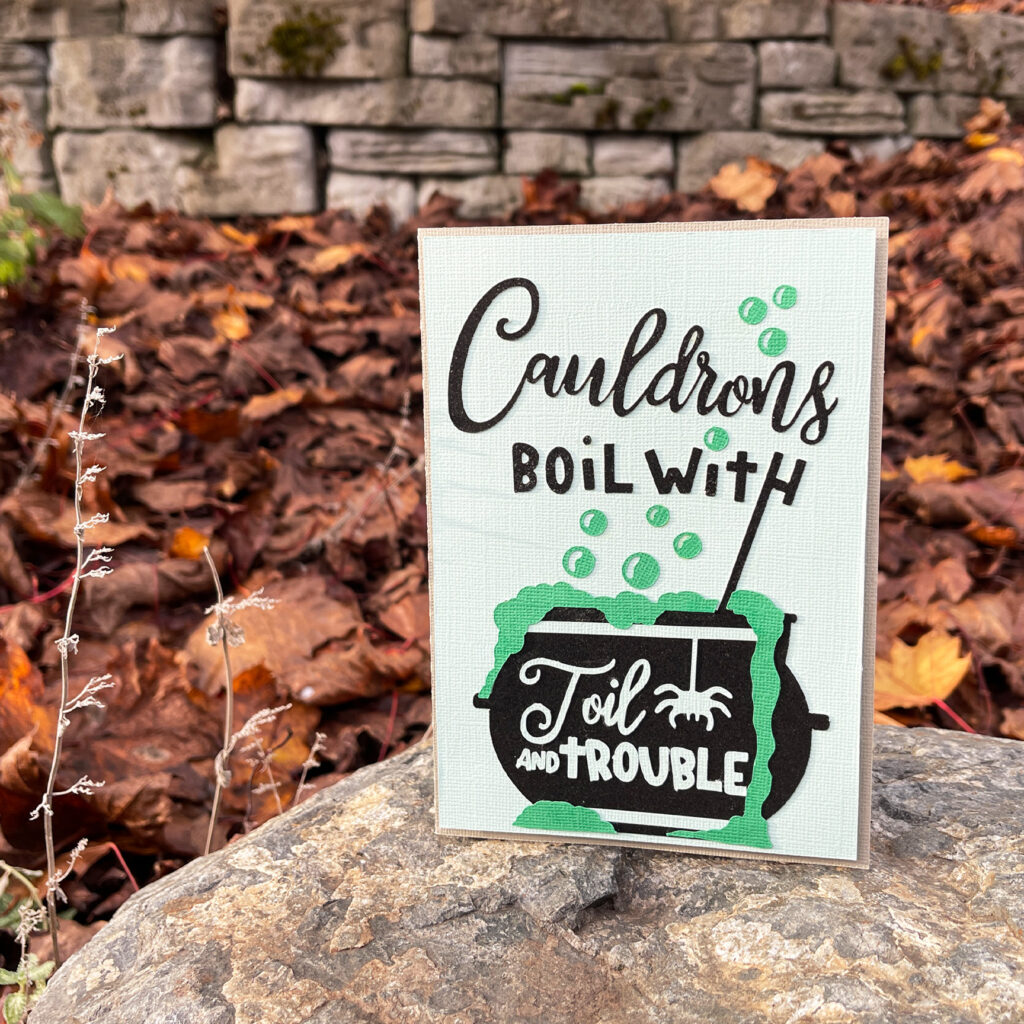 Layered paper card reading Cauldrons boil with toil and trouble featuring a bubbling cauldron and spider displayed on a rock with autumn leaves