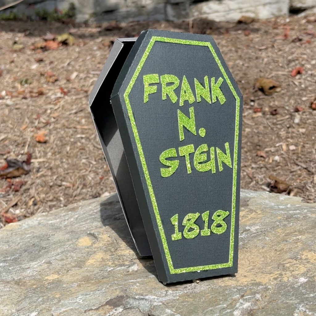Black coffin-shaped treat box with green glitter lettering reading Frank N Stein displayed on a rock outdoors