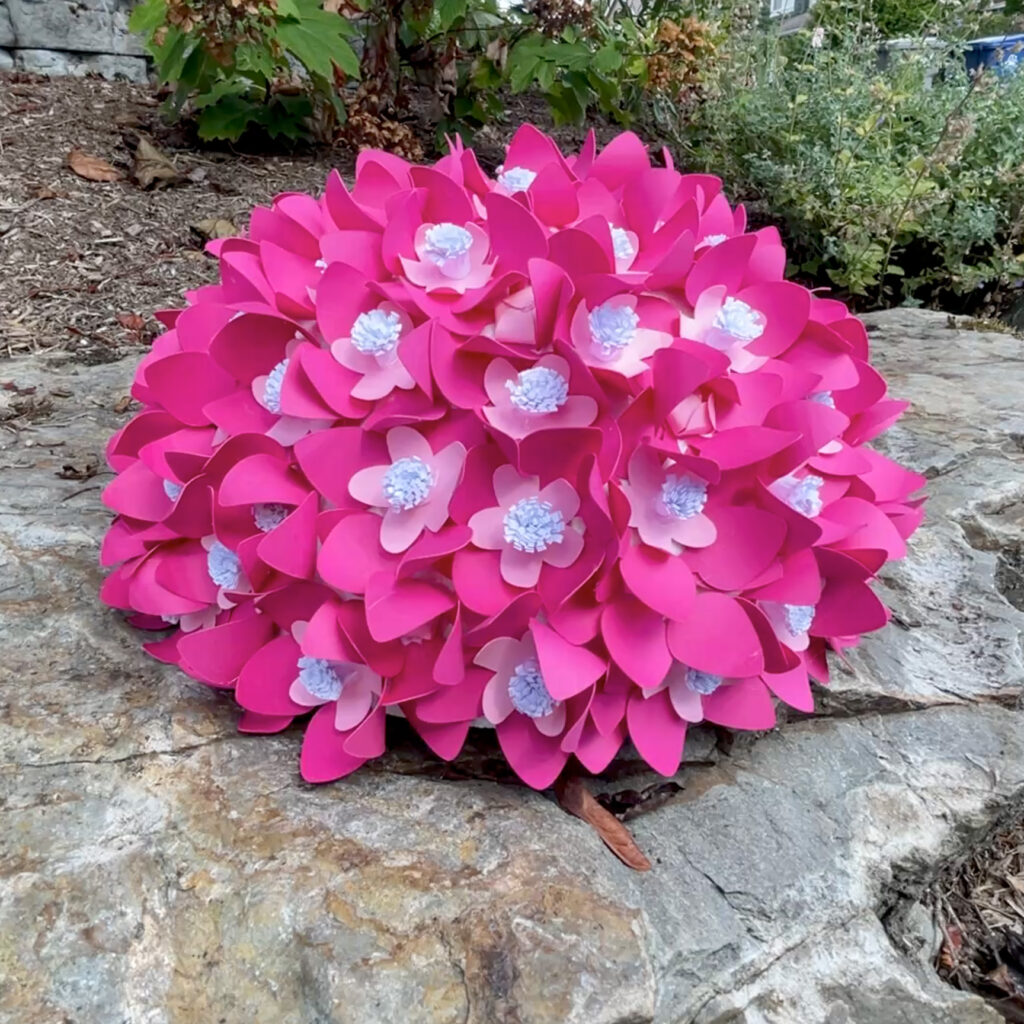 Large 3D paper hydrangea in bright pink and pale lavender tones displayed outdoors on a rock