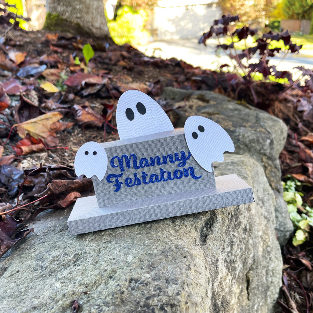 Layered paper gravestone labeled Manny Festation with three small white ghosts displayed outdoors