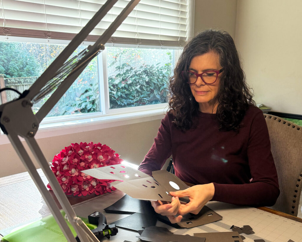 Michelle Ward assembling 3D paper craft pieces at her crafting table in natural light.