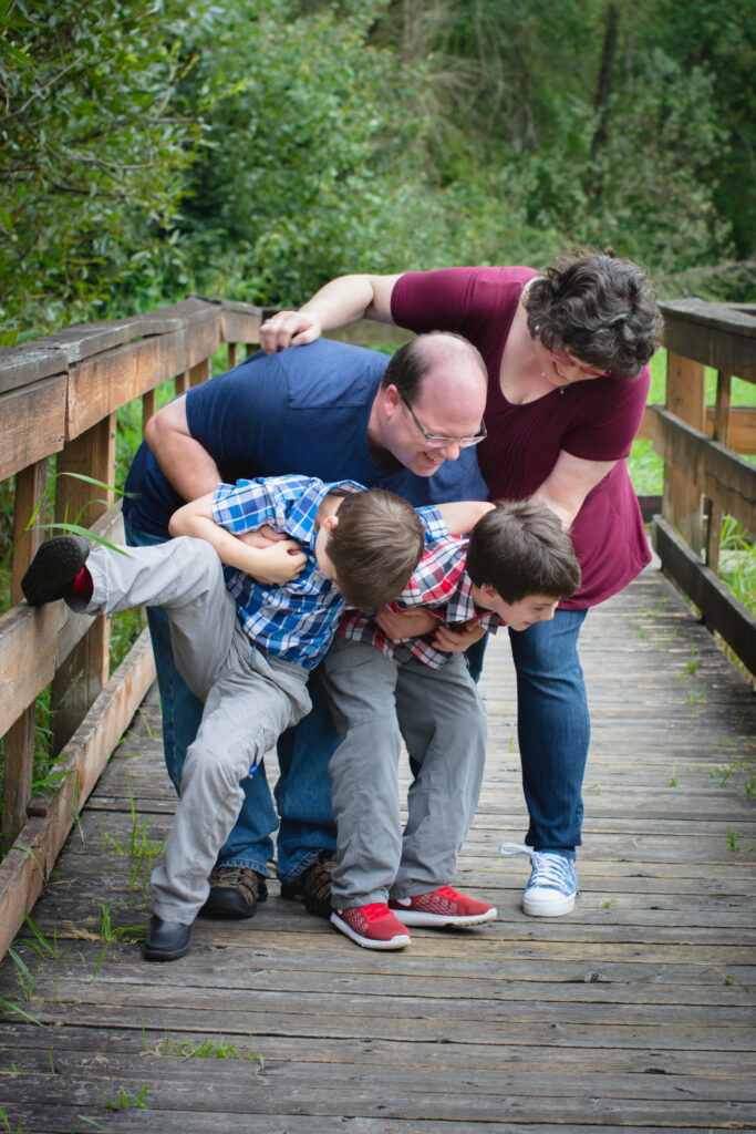 Michelle Ward laughing with her husband and two sons during a playful family moment on a wooden bridge, surrounded by green trees - a joyful snapshot of her creative, loving family.