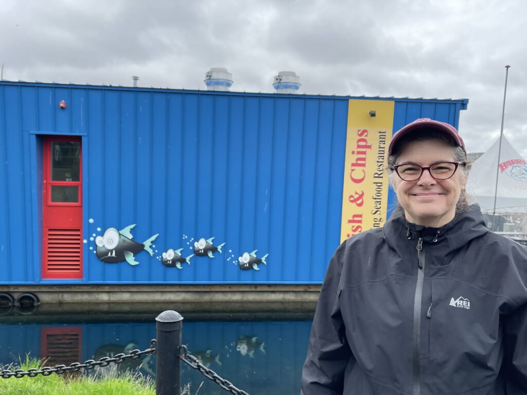 Michelle Ward smiling in front of a bright blue waterfront building in Victoria, BC, decorated with playful cartoon fish illustrations at a local fish and chips shop.
