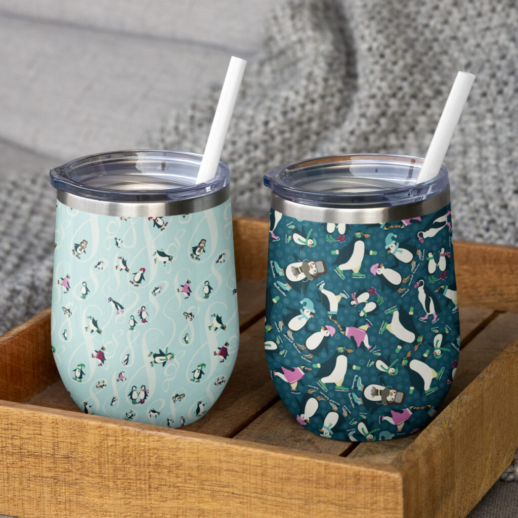 Two stainless steel wine tumblers wrapped in Waddle and Whirl repeating penguin patterns one light blue with swirls and one dark teal with penguins skates and winter accessories