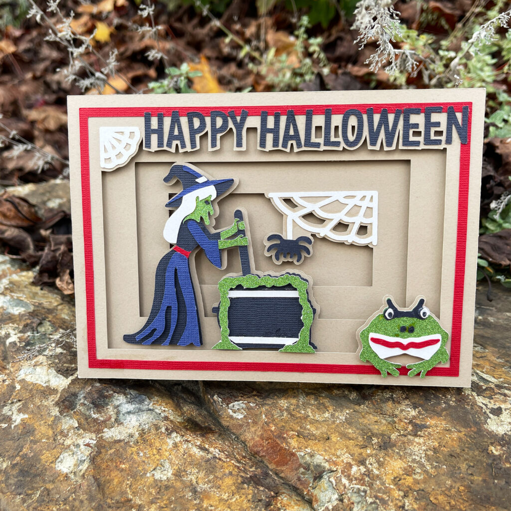 Layered Halloween shadow box card with a witch stirring a cauldron a spider web and a frog displayed on a rock outdoors