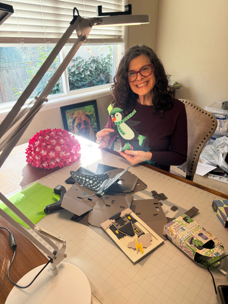 Michelle Ward holding a handmade paper penguin at her craft table, illustrating what belonging really means through creativity and cozy chaos.