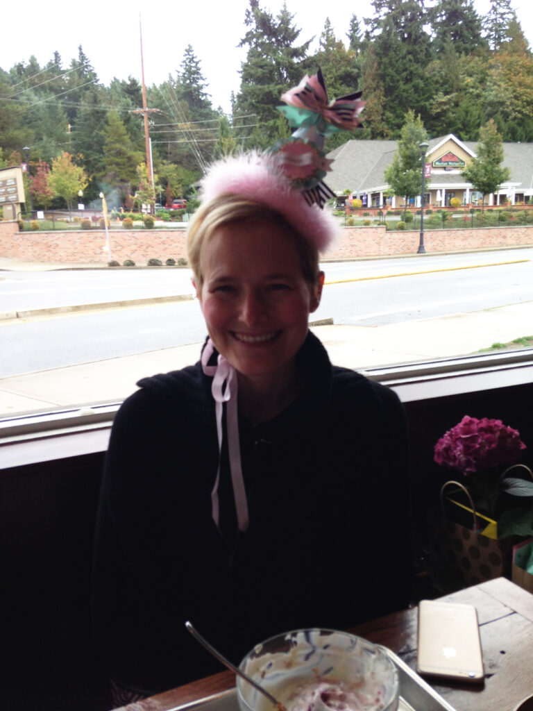 Molly smiling in a café while wearing a pink fluffy headpiece with ribbons and bows.